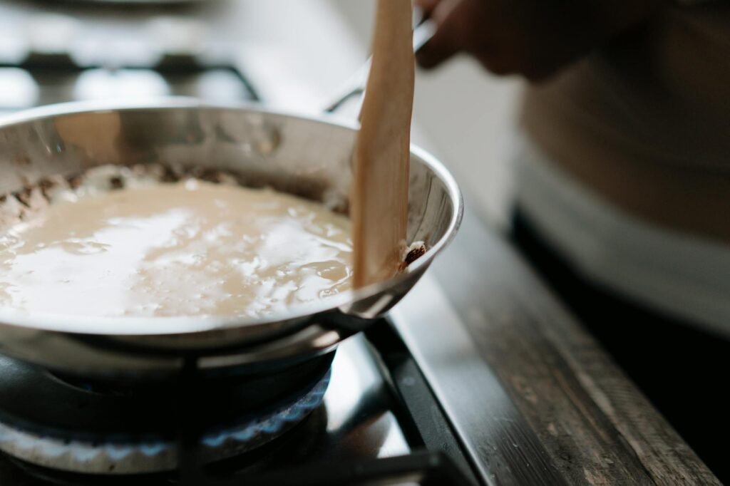 A cook whisking homemade gravy in a saucepan beside a roasting pan with browned drippings.